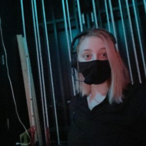 A dimly lit image of Hayley Bamford CSOPA Stage Manager, standing in front of a fly galley. She is wearing all black clothes for backstage, including a black facemask. she is also wearing a theatre headset.