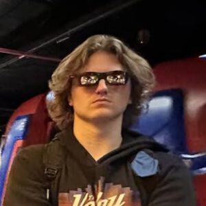 Picture of Noah Buhler, JR Stage Stage Manager. He has long blond hair and rectangular sunglasses. He is staring unsmilingly at the camera.