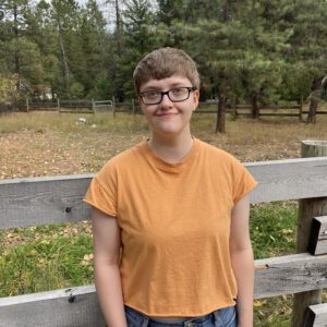 An Image of Hailey Bate, Devised Stage Manager. They are outside by a wood fence, softly smiling. They wear a yellow Tshirt, closely cropped blonde hair, and black, rectangular glasses. 
